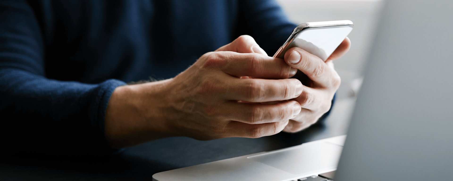 A closeup of a man sitting in front of a laptop holding and using a mobile phone to contact technical support